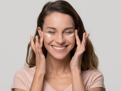 femme aux cheveux bruns qui sourit et qui se tire les yeux en application de la crème