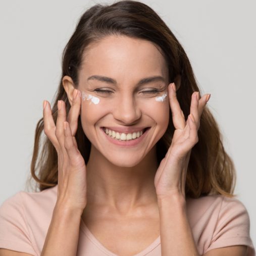 femme aux cheveux bruns qui sourit et qui se tire les yeux en application de la crème